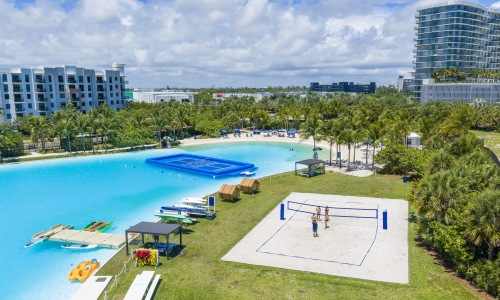 a body of water in front of trees and buildings with a sand volley ball court